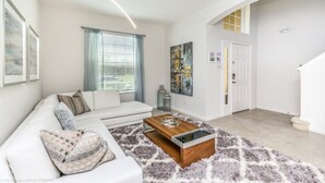 Un salón moderno con un sofá blanco, una mesa de centro de madera y una alfombra mullida. Hay una puerta con un panel de vidrio y un cuadro en la pared.