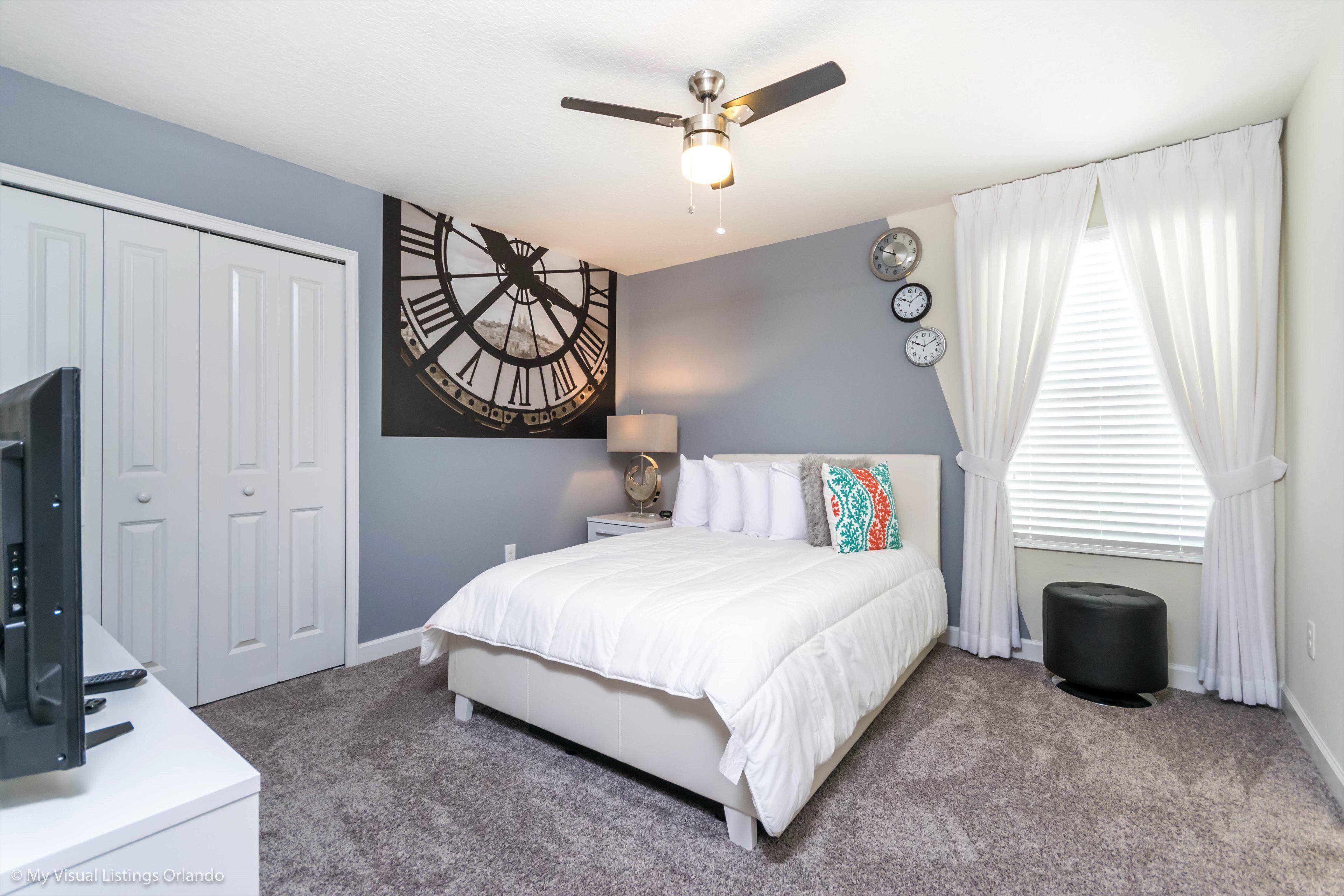 A bedroom with a bed, a ceiling fan, a clock, and a window with curtains.