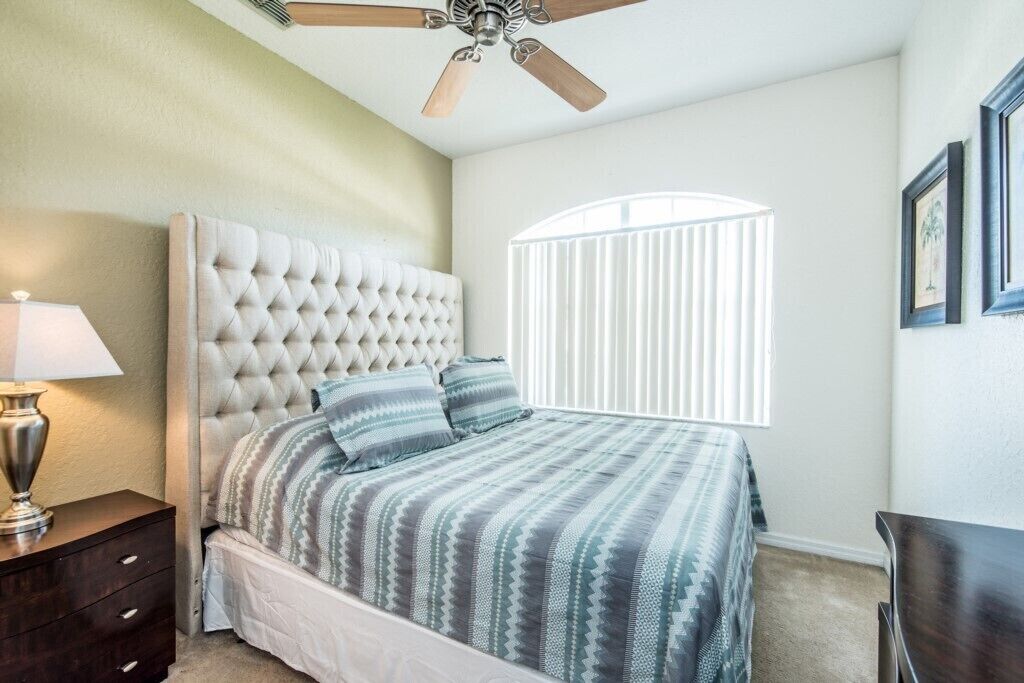 Une chambre à coucher avec une tête de lit capitonnée, un lit avec une literie à rayures, un ventilateur de plafond, une table de chevet, une lampe, une fenêtre avec des stores et des tableaux encadrés accrochés au mur.