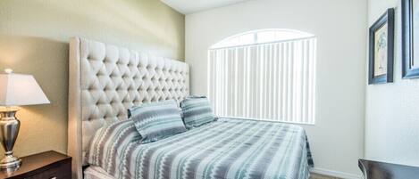 Une chambre à coucher avec une tête de lit capitonnée, un lit avec une literie à rayures, un ventilateur de plafond, une table de chevet, une lampe, une fenêtre avec des stores et des tableaux encadrés accrochés au mur.