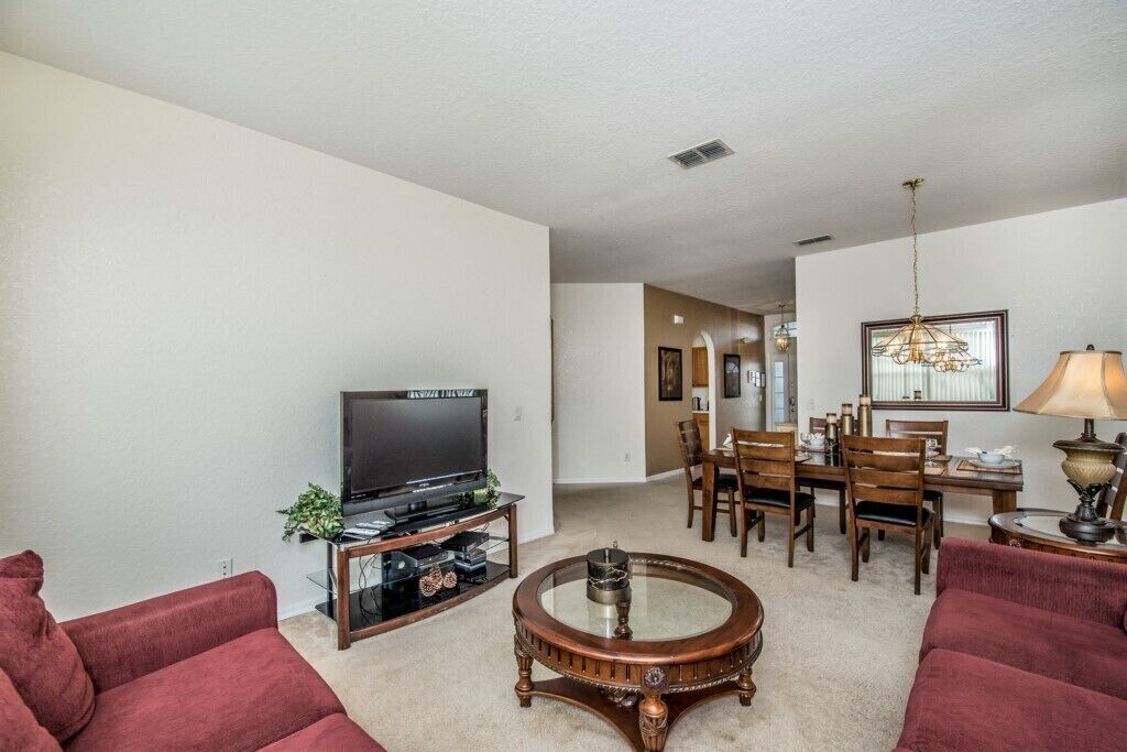 A living room with a flat-screen TV, a coffee table, and red sofas. There is a dining area with a table and chairs.