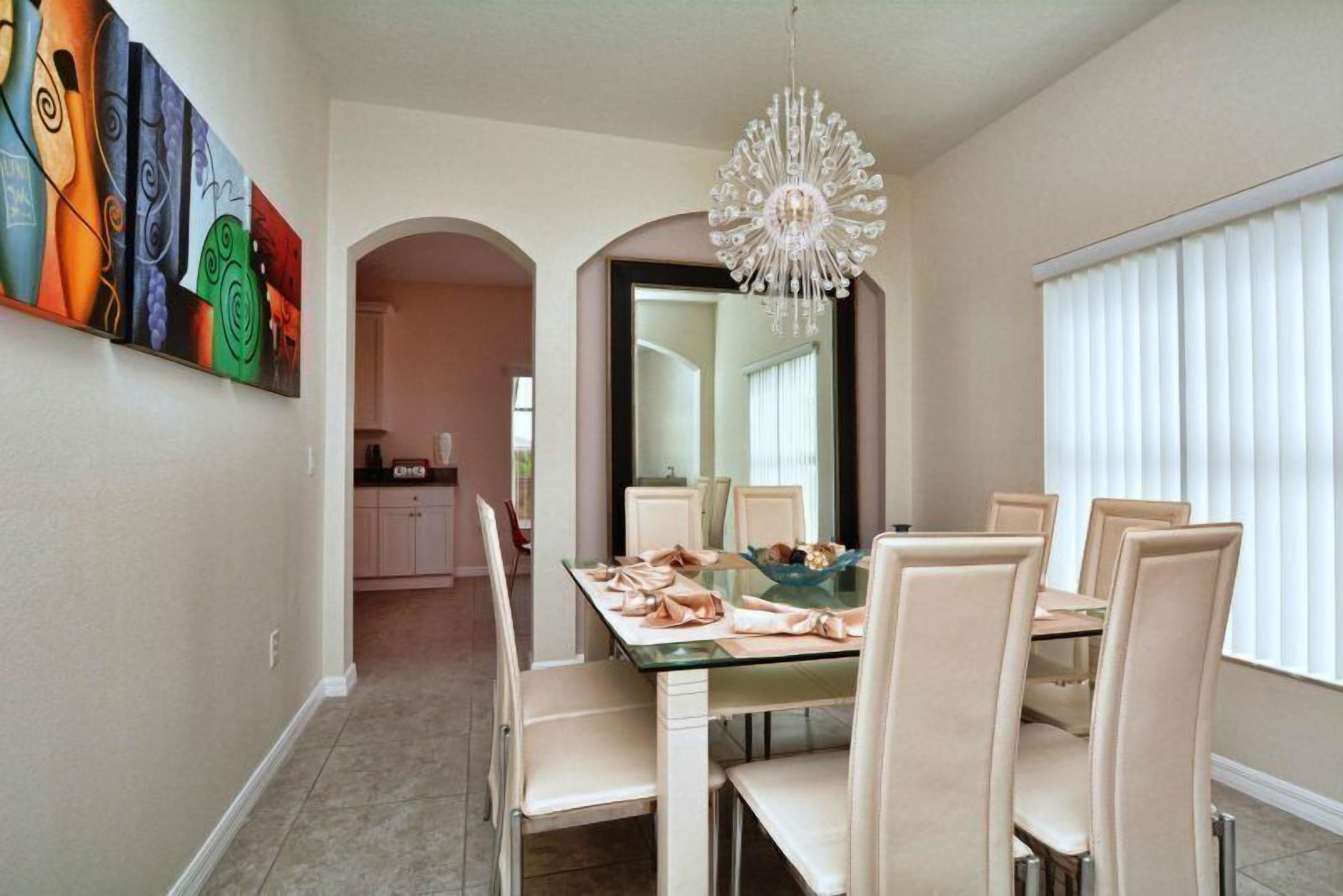 A dining area with a glass table, white chairs, and a chandelier. There is a colorful abstract painting on the wall and a kitchen in the background.