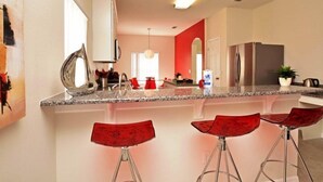 A modern kitchen with a granite countertop, red bar stools, a stainless steel refrigerator, and a pendant light. - 616bd (Davenport)