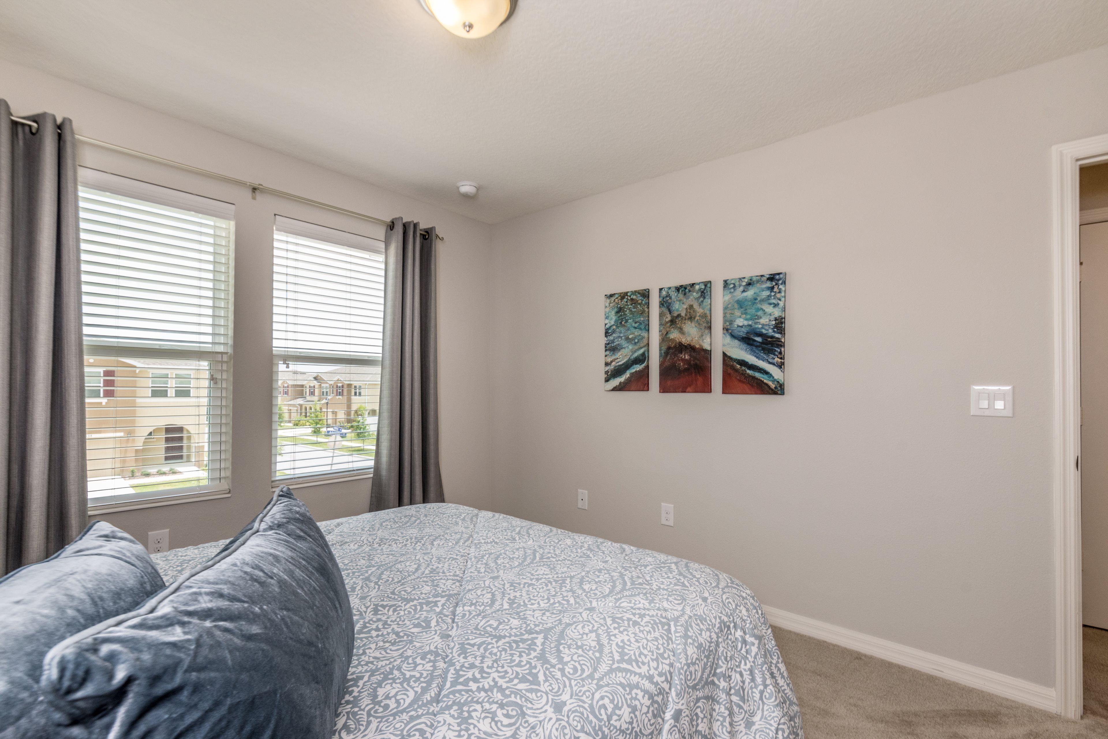 A bedroom with a large bed, two windows with blinds, and three wall-mounted paintings.