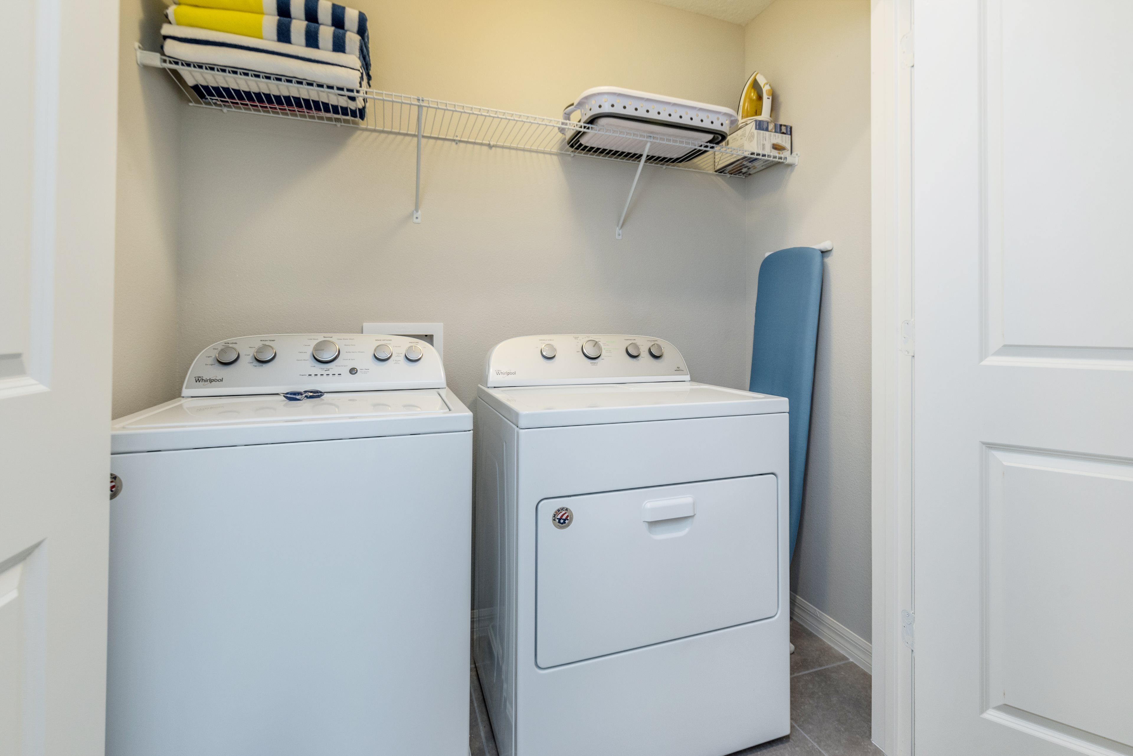 Een wasserette met twee witte Whirlpool wasmachines en drogers, een metalen plank met opgevouwen handdoeken, en een blauwe mat op de vloer.