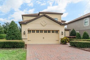 Une maison beige à deux étages, dotée d’un garage, entourée de haies bien entretenues et bordée d’un chemin en briques.