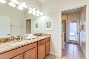 Une salle de bain dotée d’un comptoir en granit, d’un lavabo double et d’un grand miroir. Des tableaux encadrés ornent le mur, et un porte-serviettes contient des serviettes.