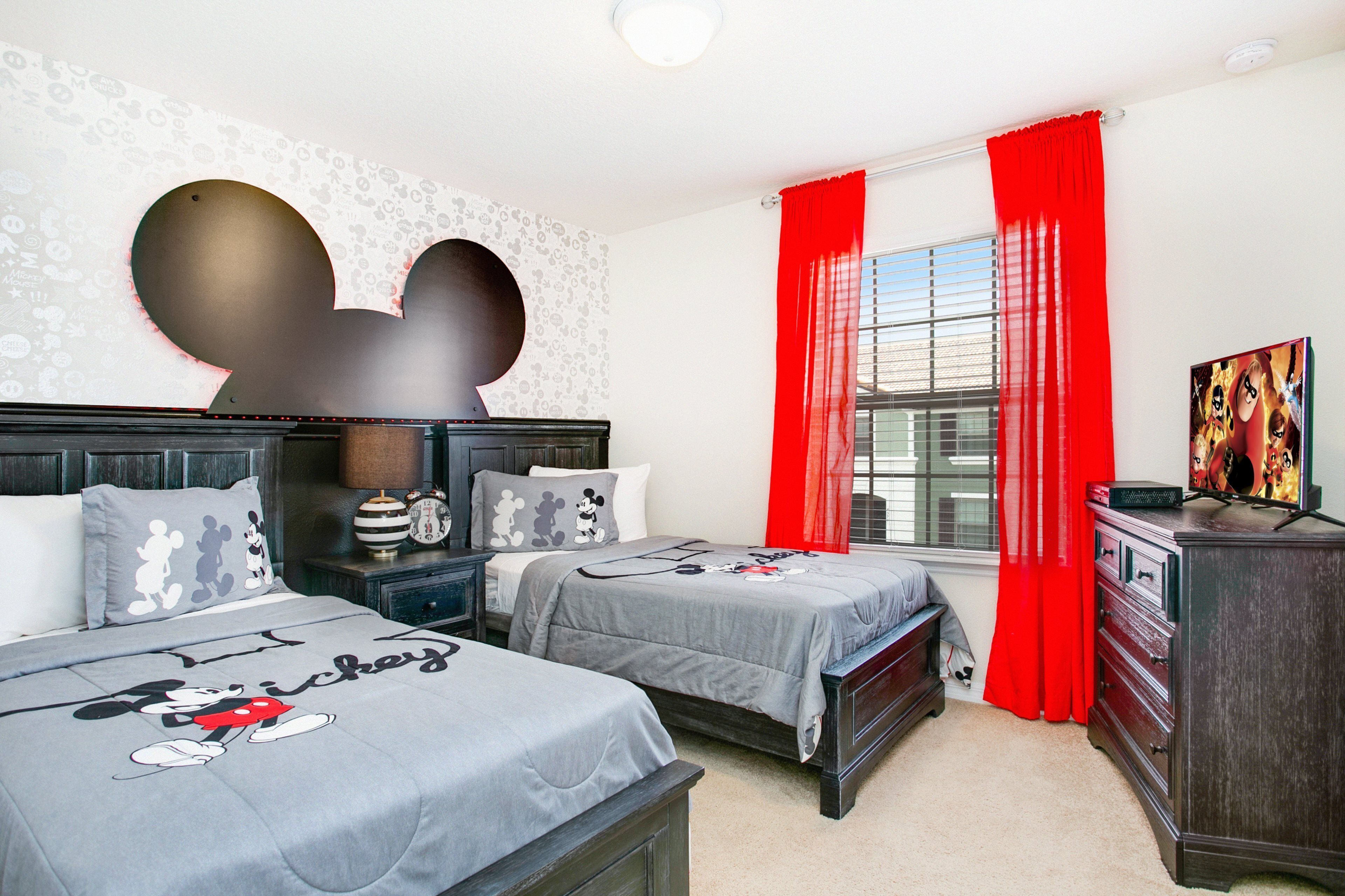 Ein Schlafzimmer mit zwei Betten, einer Wanddekoration mit Mickey Mouse, einem Schminktisch und einem Fenster mit roten Vorhängen.