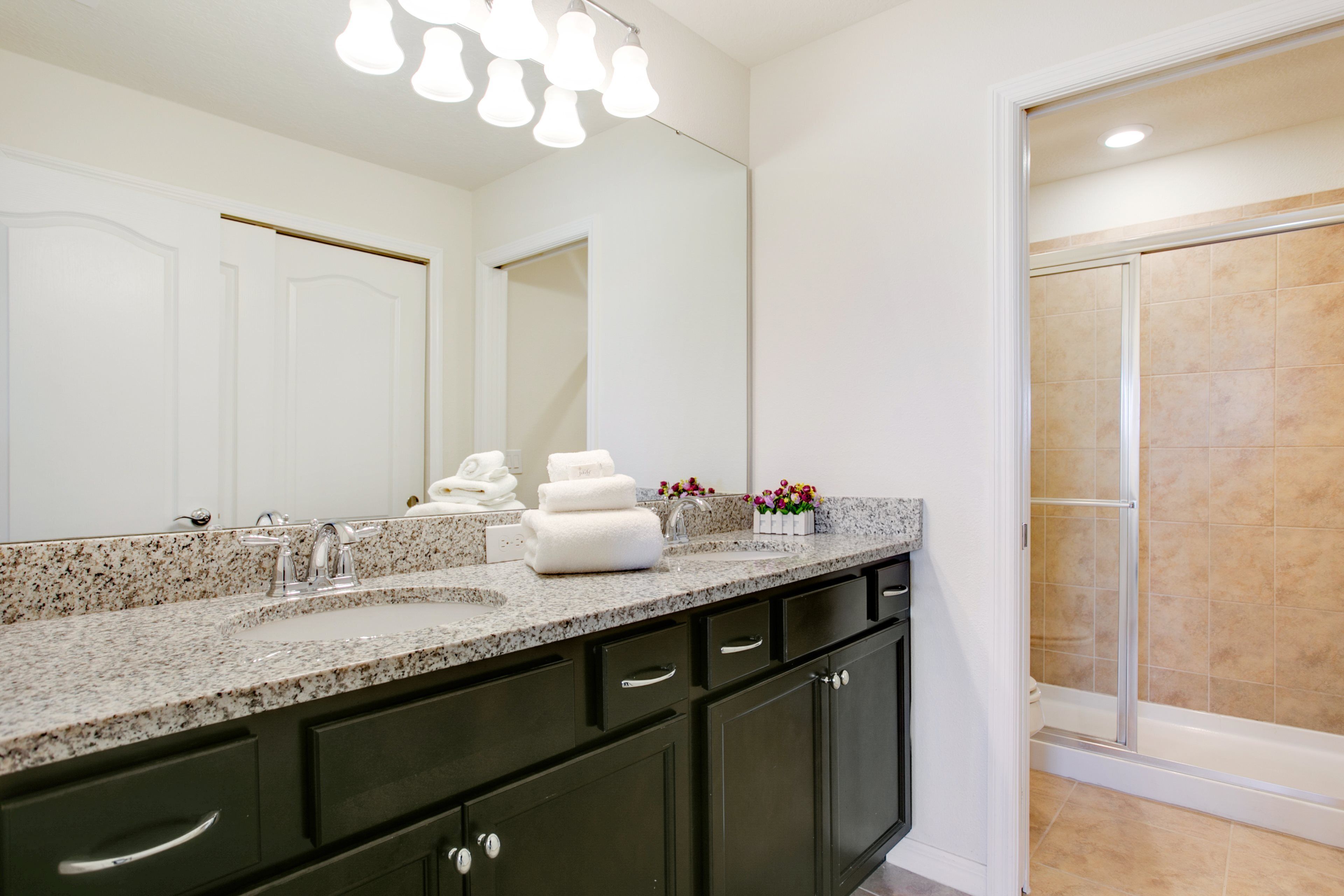 A modern bathroom with a granite countertop, dark cabinets, a large mirror, and a walk-in shower.