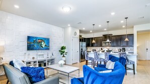 Ein modernes Wohnzimmer mit grauem Sofa und blauen Sesseln, einem Flachbildfernseher an einer weißen Ziegelwand und einer Küchenzeile mit dunklen Schränken und Pendelleuchten.