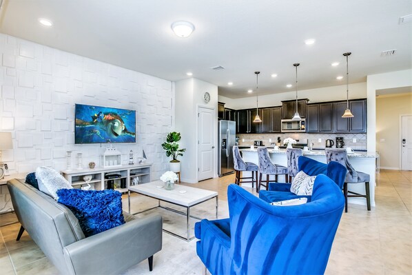 A modern living room with a grey sofa and blue armchairs, a flat-screen TV mounted on a white brick wall, and a kitchen area with dark cabinets and pendant lighting.