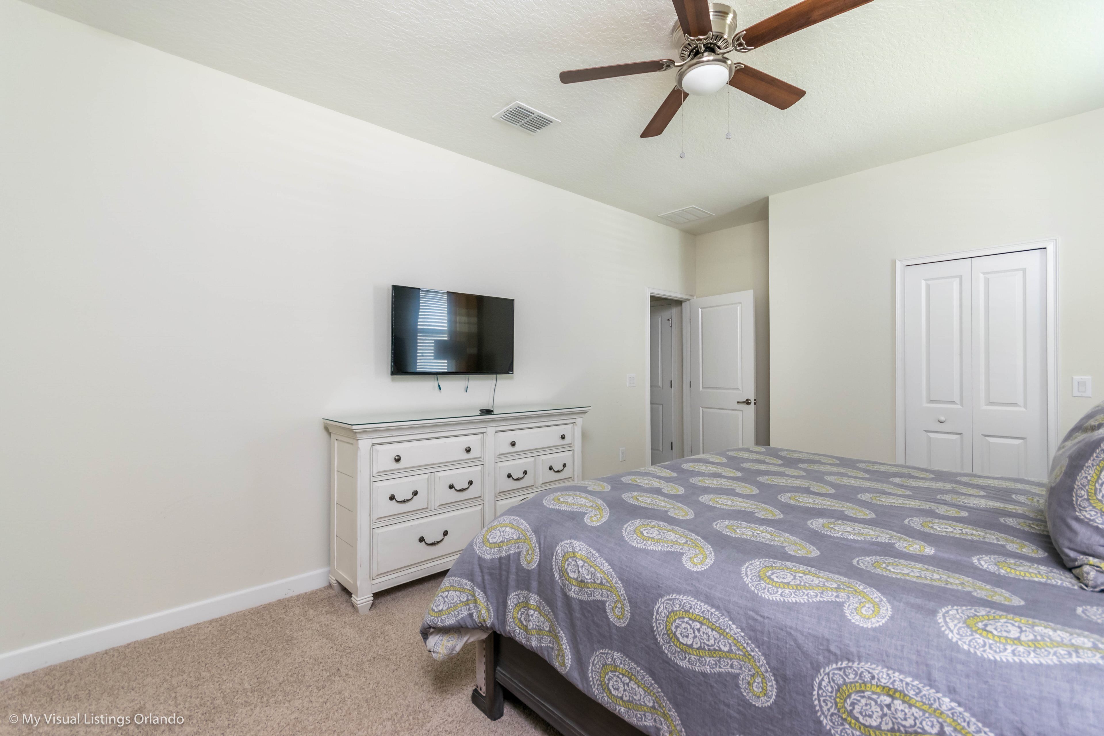 A bedroom with a bed, a dresser, a ceiling fan, a television, and a door.