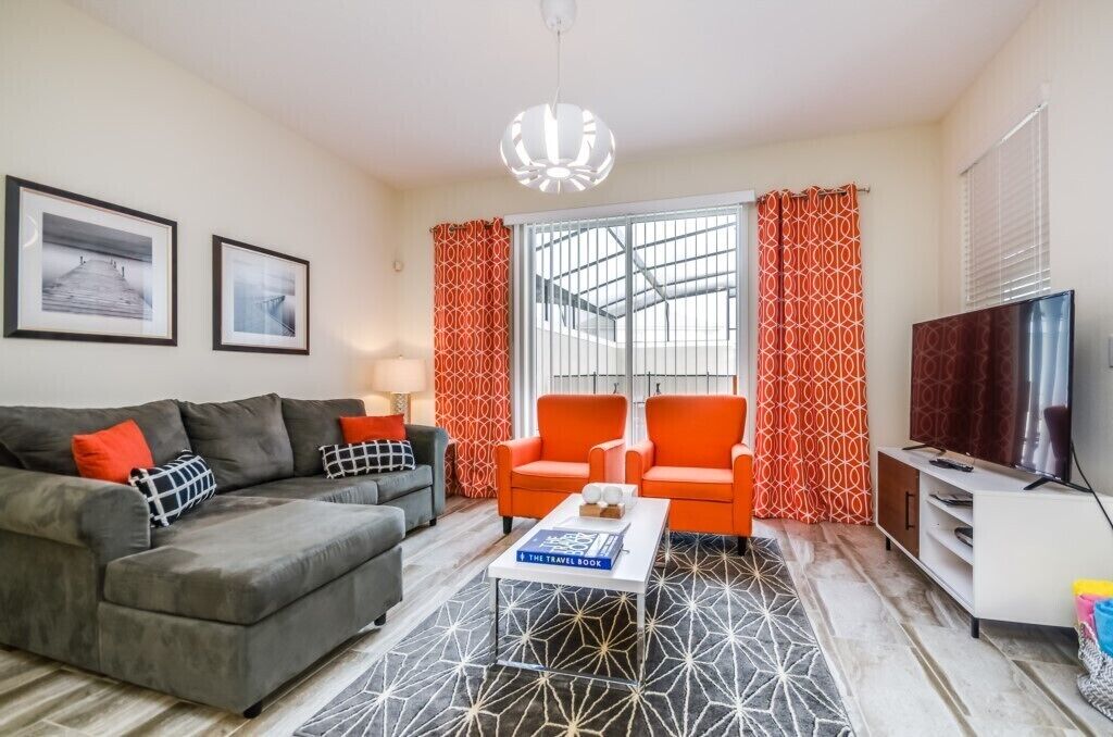 A living room with a grey sofa, orange armchairs, a coffee table with a book, a TV, and patterned curtains.