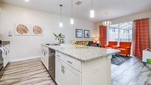 Eine moderne Küche mit weißen Schränken, Granit Arbeitsplatten und Pendelleuchten. Ein Essbereich mit orangefarbenen Stühlen und einem gemusterten Teppich.