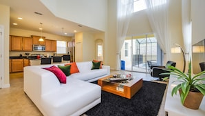 A spacious living area with a white sofa, black rug, and a wooden coffee table. There is a kitchen in the background with modern appliances and a dining area with black chairs. - Fantastic Resort 5 Bed 5 Bath Villa With Pool (Kissimmee)