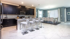 A modern kitchen with dark cabinets, a central island with bar stools, a dining area with a table and chairs, and a large window with curtains.