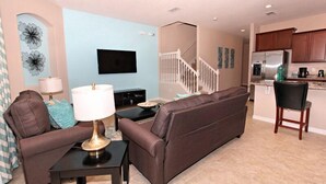 A living room with a sofa, a coffee table, and a TV. There is a staircase leading to the upper floor. - Townhome with Pool at Solterra (Davenport)