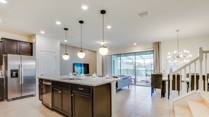 A modern kitchen with a large island, stainless steel appliances, pendant lighting, and a view to a dining area with a staircase.