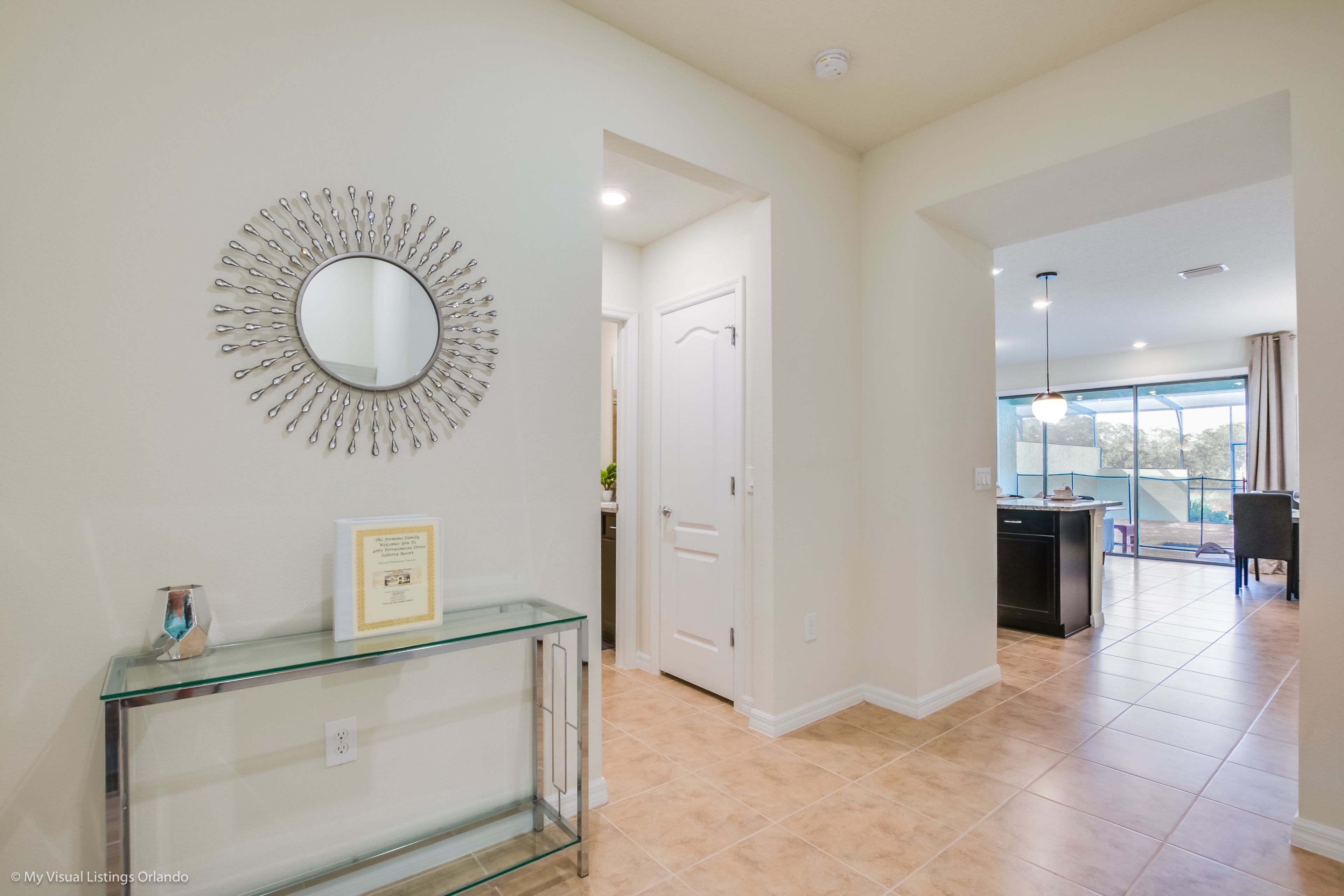 A modern interior with a glass console table, a decorative mirror, and a kitchen area visible through an open door.