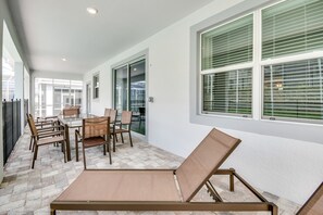 Eine sonnendurchflutete Terrasse mit Esstisch und Stühlen, einem Liegestuhl und großen Fenstern mit Jalousien.