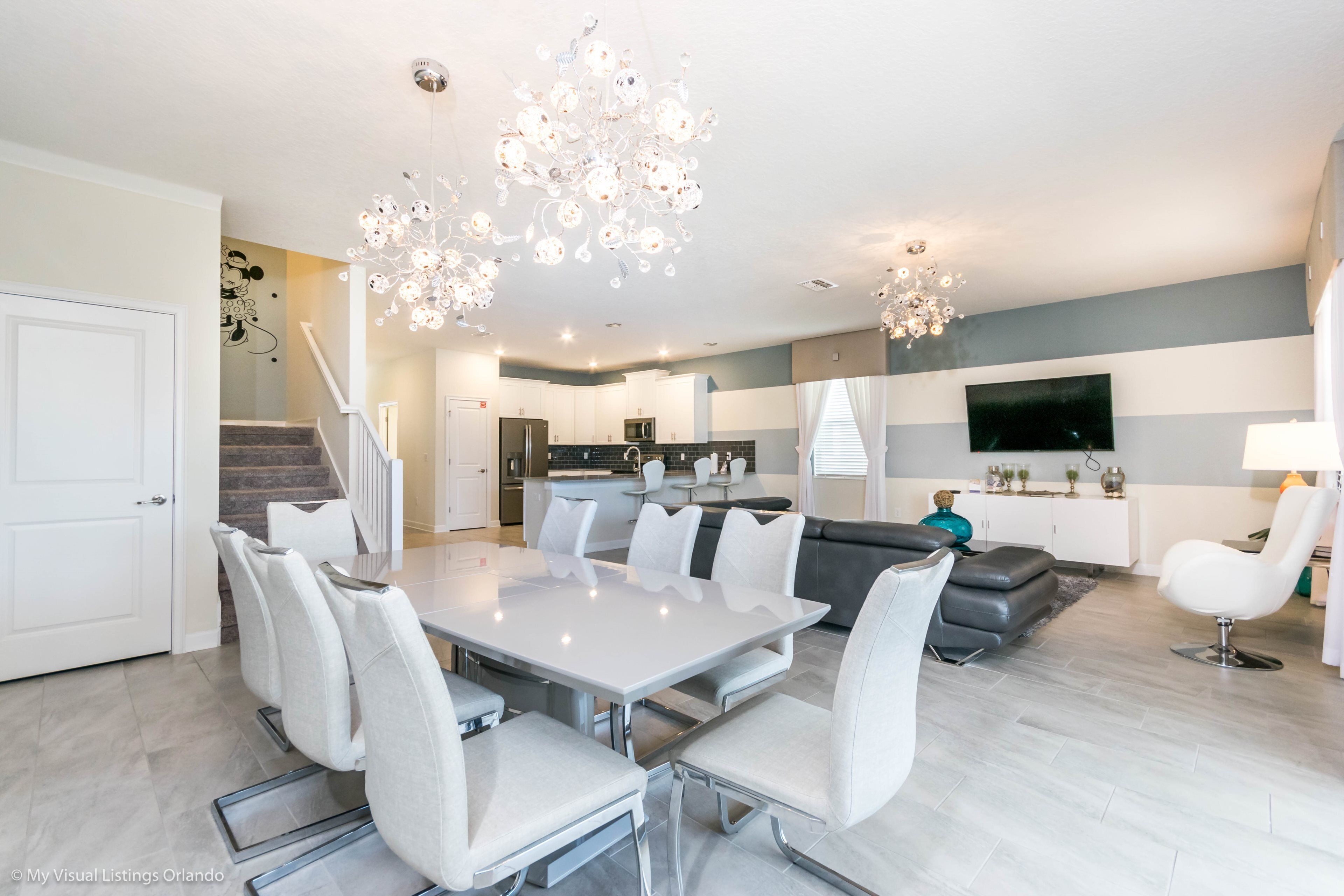 Eine moderne Küche mit Einbauofen, ein Essbereich mit weißem Tisch und Stühlen sowie ein Wohnbereich mit grauem Sofa und Wand-Fernseher.