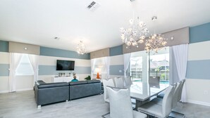 A modern living room with a grey sofa, a dining table with chairs, a TV, and a chandelier. - 1686 Champions Gate 8 Bedroom Resort Villa (Davenport)