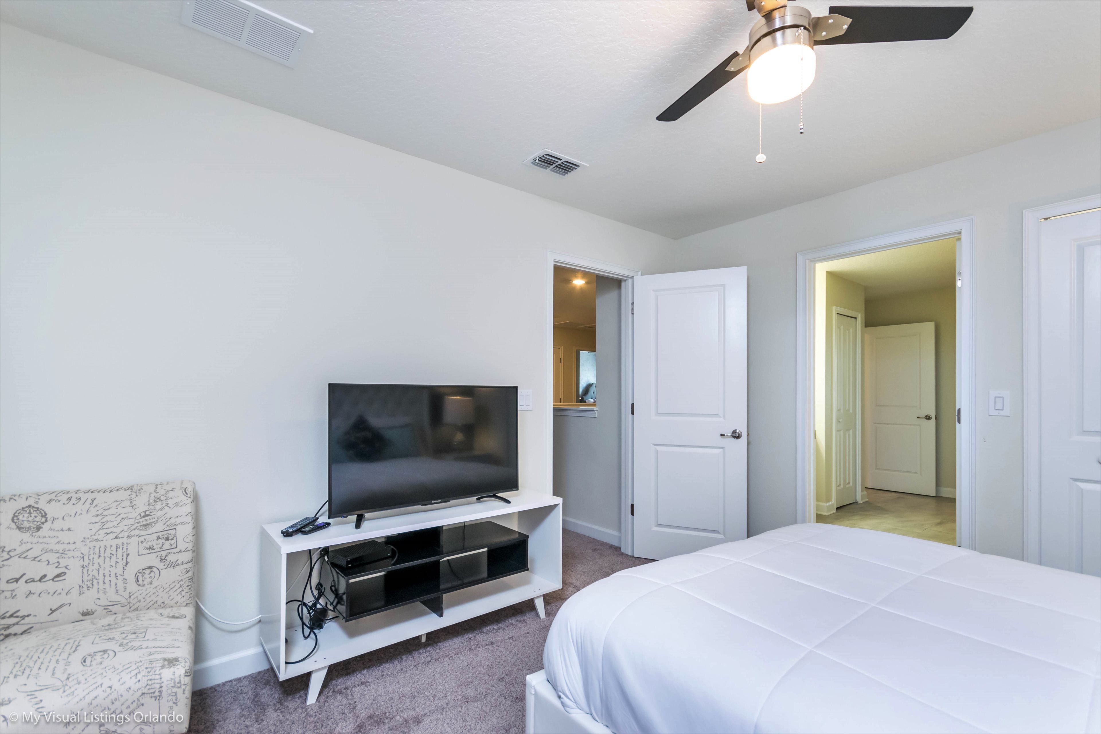 A bedroom with a bed, a TV on a white entertainment unit, a ceiling fan, and white doors leading to other rooms.
