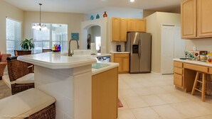 A modern kitchen with a central island, stainless steel refrigerator, wooden cabinets, and a dining area with wicker chairs. - Fantastically Decorated 4 bed 3 Bath (Davenport)