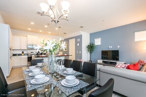 A modern kitchen with white cabinets, a dining area with a glass table set for six, and a living area with a sofa and TV.