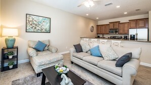 A living room with a sofa, ottoman, and coffee table, a ceiling fan, and a kitchen in the background. - 4 Bedroom 3 Bathroom Town Home With Pool (Kissimmee)