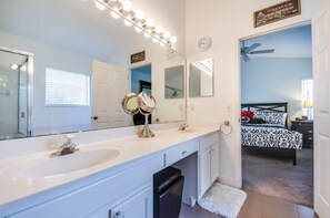 A bathroom with a double sink, vanity, and a large mirror with lights. There is a bed with a black and white patterned bedspread in the adjacent room. - 633tc Modern 3 Bedroom Villa (Davenport)