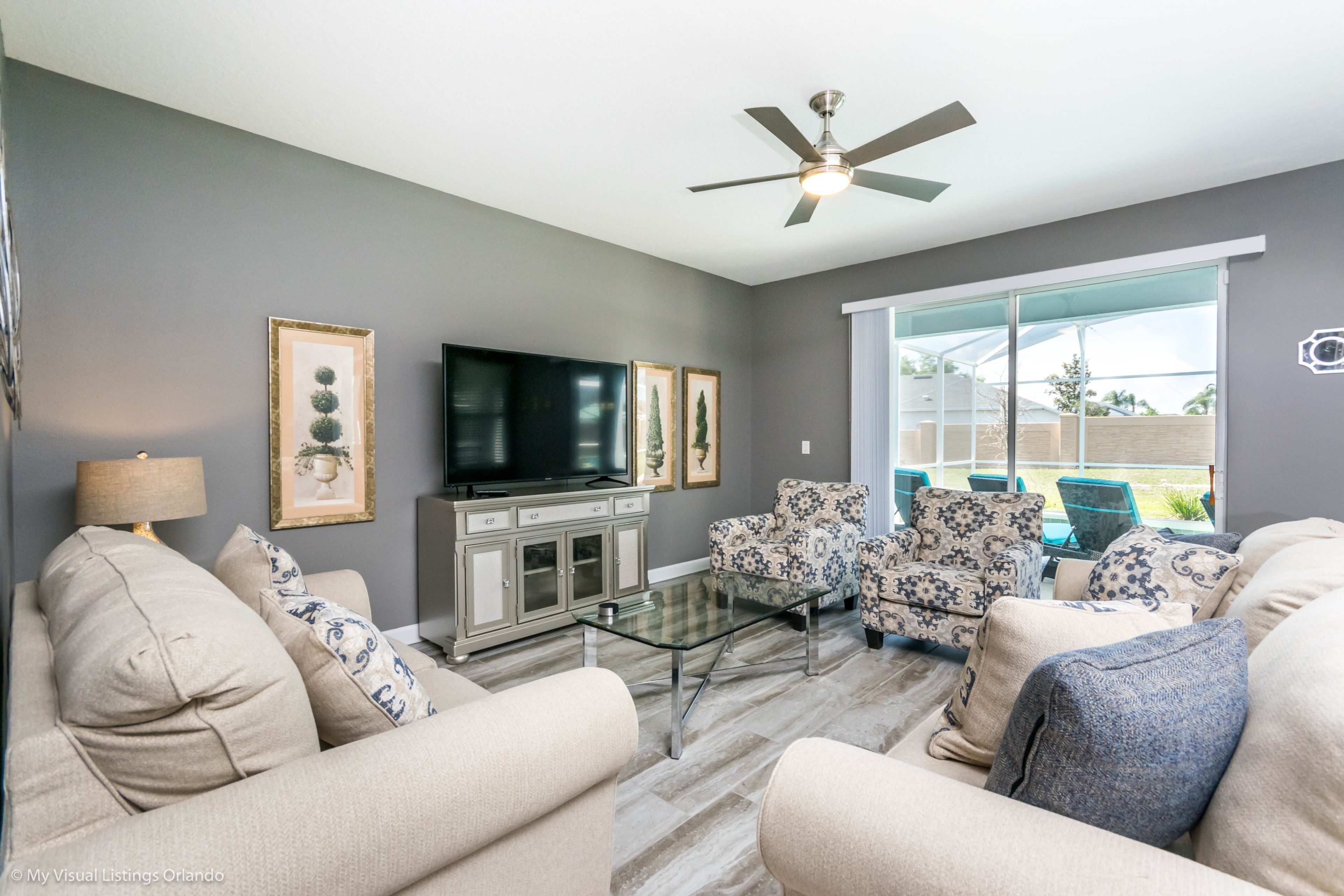 A modern living room with a flat-screen TV, patterned sofas, a glass coffee table, and a ceiling fan.