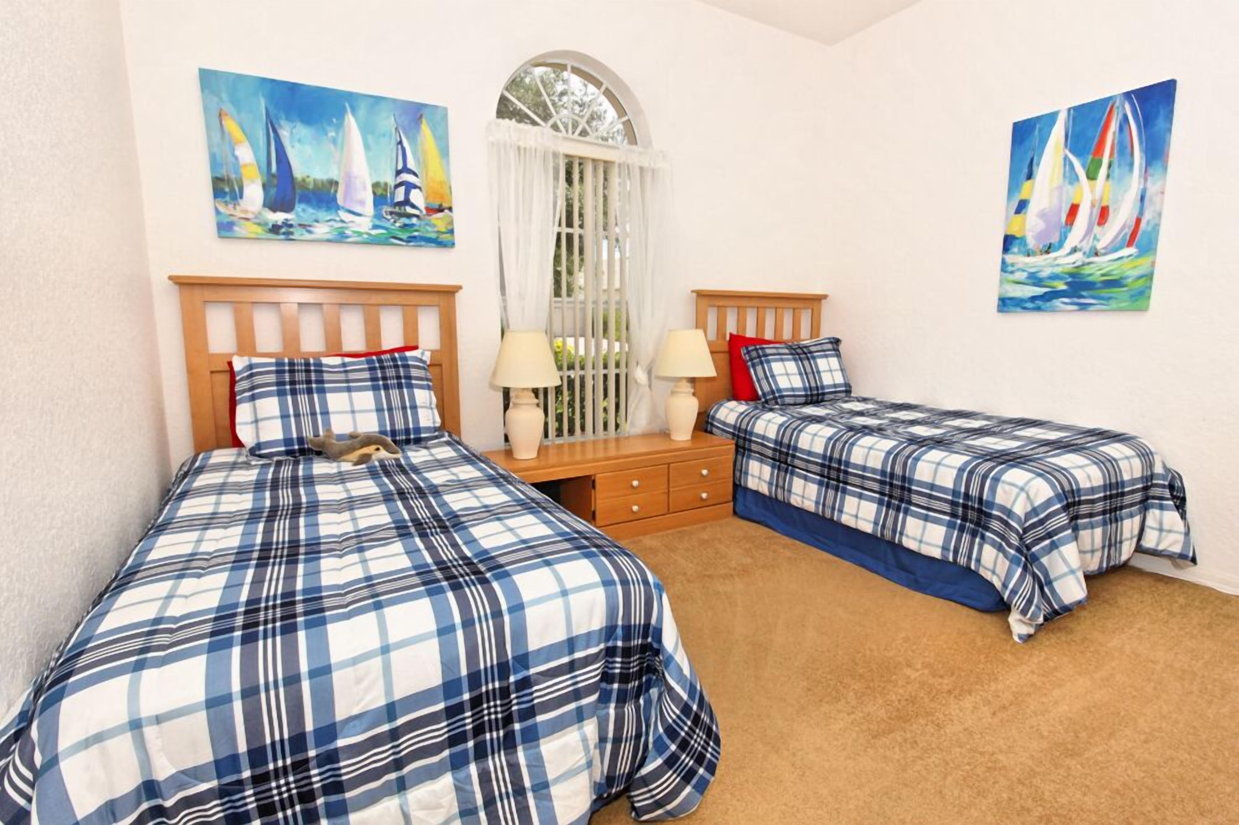 A bedroom with two beds, each with a blue and white checkered bedspread, a wooden headboard, and a nightstand with a lamp. There are two paintings of sailboats on the walls.