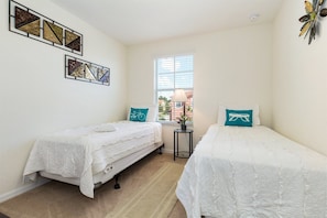 Um quarto pequeno e iluminado com duas camas de solteiro, uma janela com persianas e quadros nas paredes.