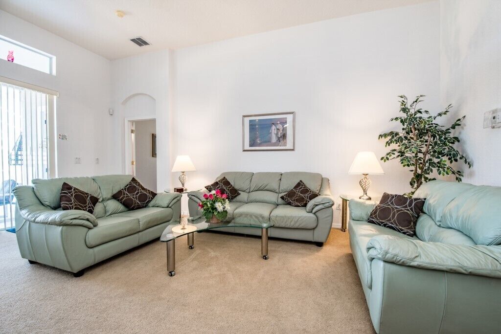 A living room with two green sofas, a glass coffee table, and two lamps. There is a framed picture on the wall and a potted plant.