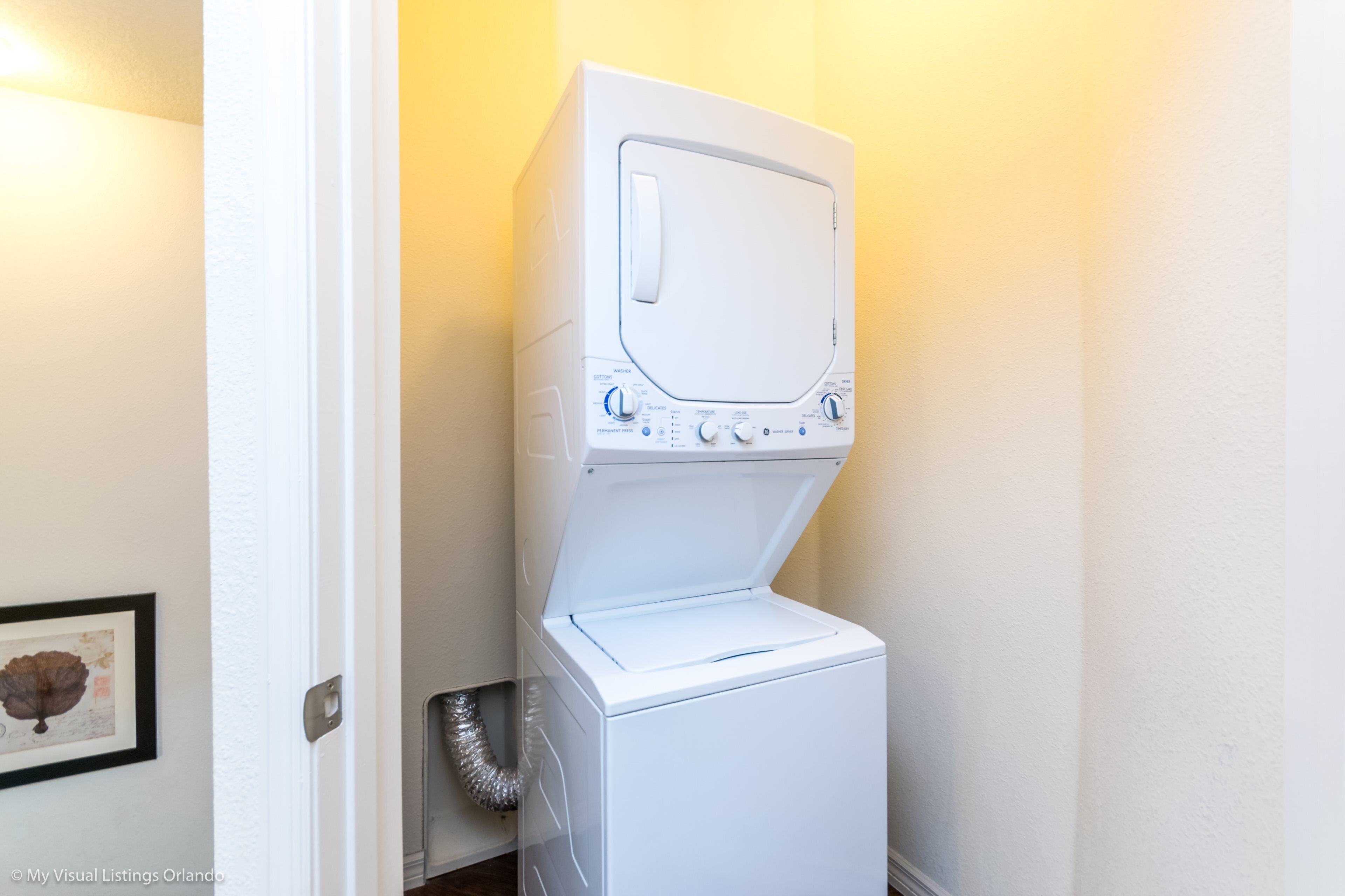 A compact, white stacked washer and dryer unit in a small laundry room with a framed picture on the wall.