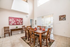 A dining area with a wooden table set for four, surrounded by six chairs. There is a large window with blinds, a door leading to another room, and a wall-mounted painting.