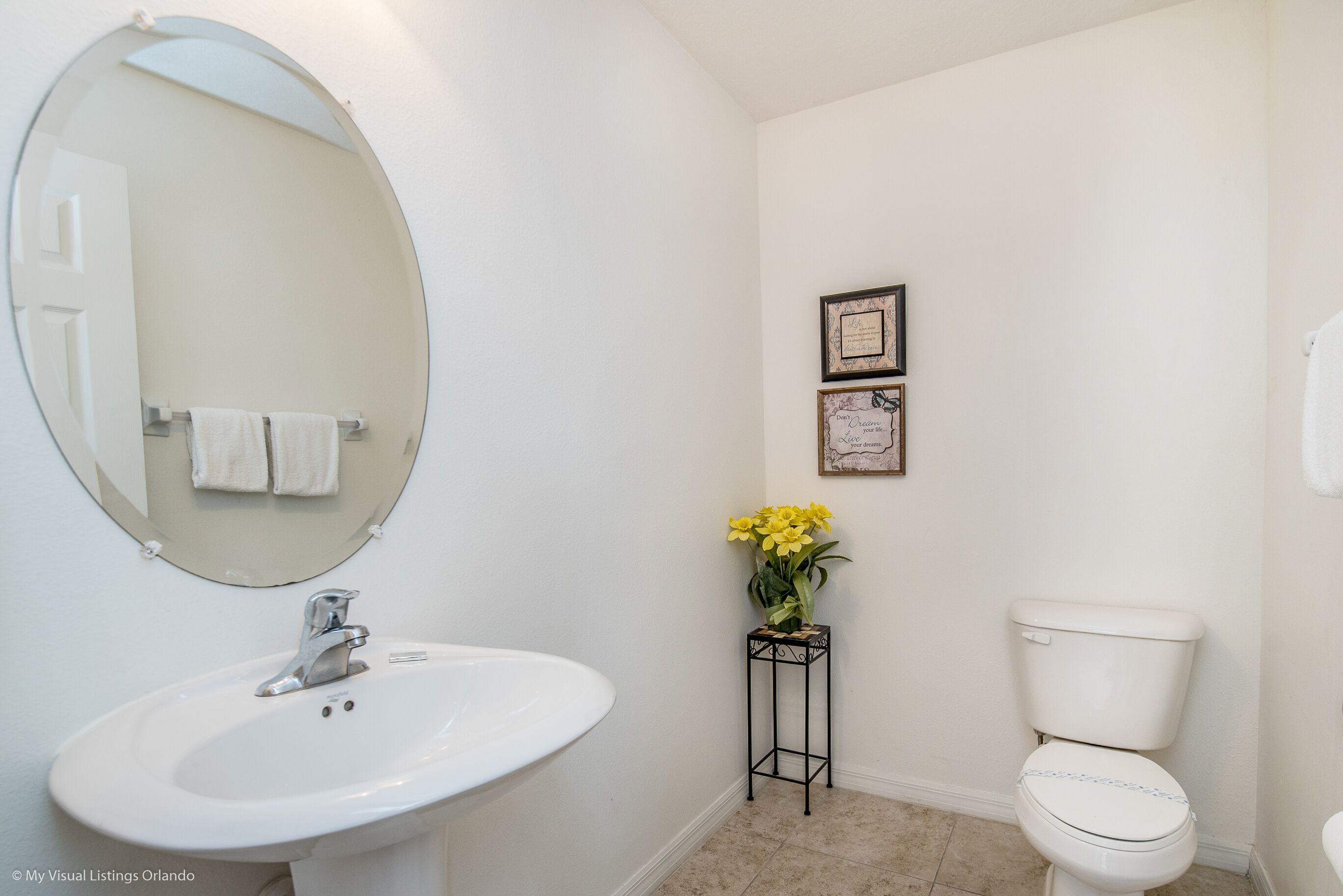 A bathroom with a white sink, a round mirror, a toilet, and a small table with a vase of yellow flowers.