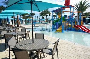 Zona de piscina con sillones de descanso y sombrillas, un tobogán de agua y una estructura de juegos.