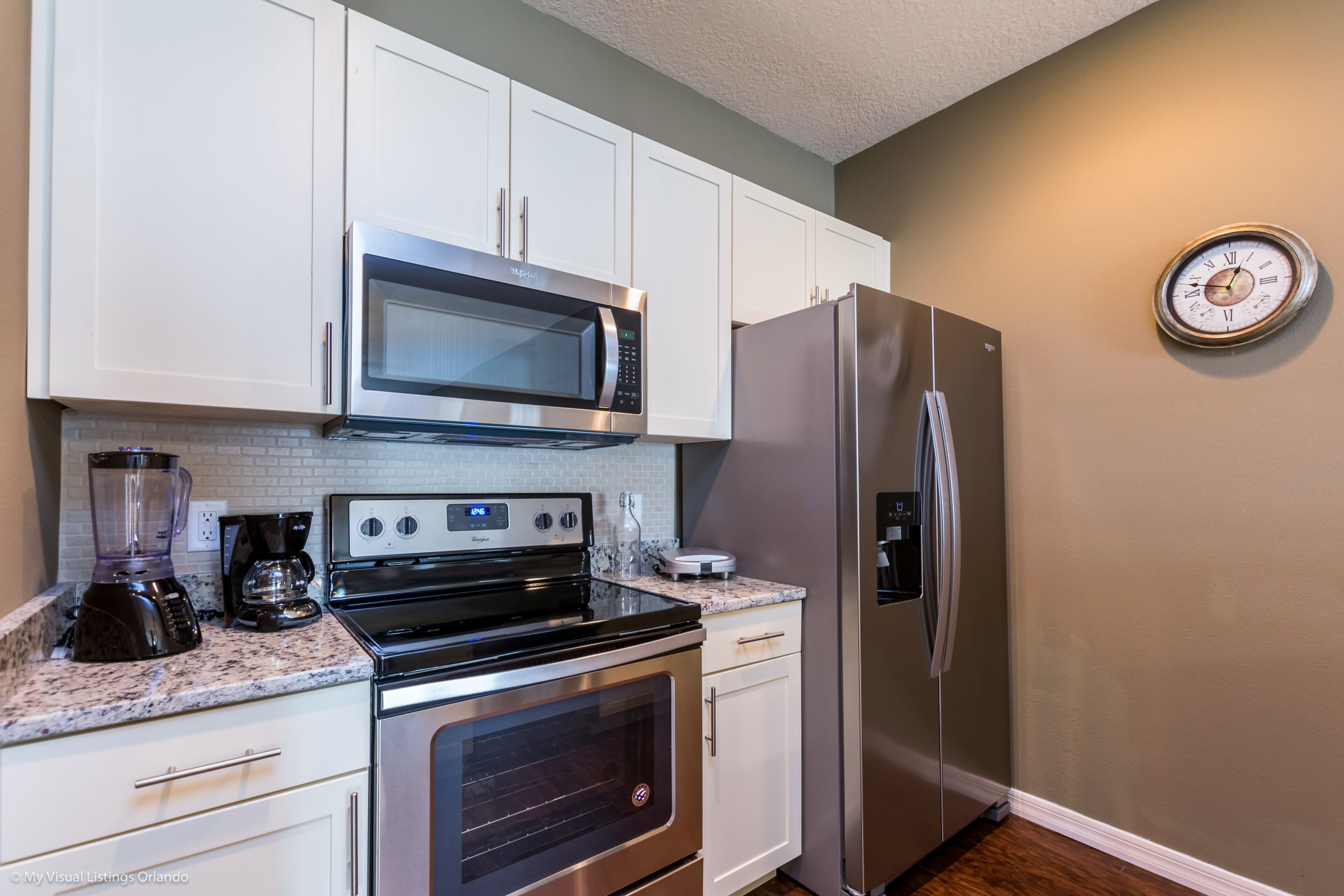 Una cocina con armarios blancos, un refrigerador de acero inoxidable, un microondas, un horno y un reloj en la pared.