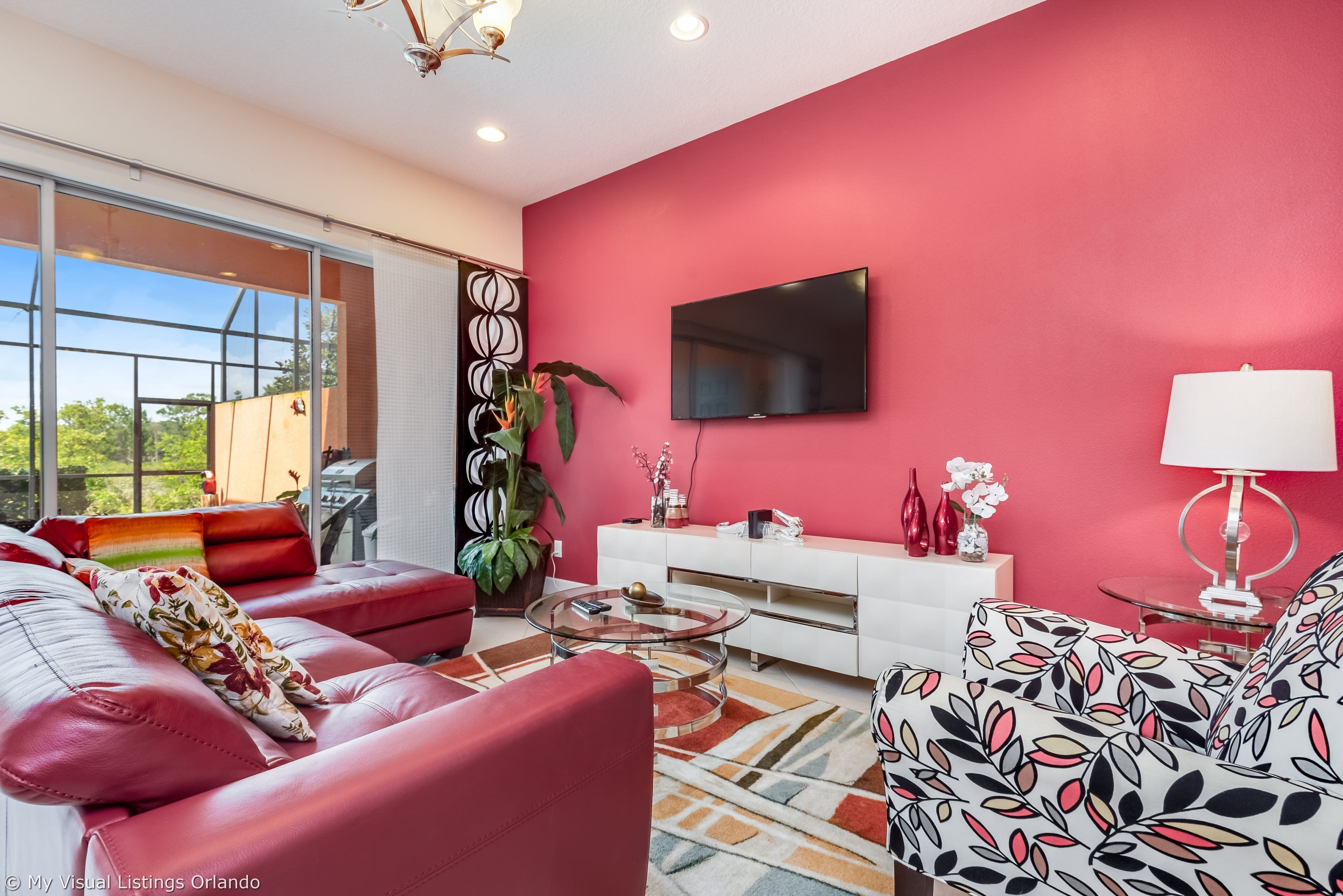 A modern living room with a red accent wall, a flat-screen TV, a glass coffee table, and a sofa with floral cushions.