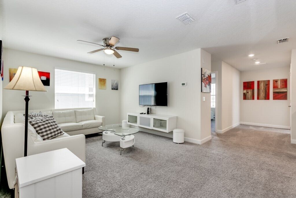 A modern living room with a white sofa, a glass coffee table, a ceiling fan, and a flat-screen TV on the wall.