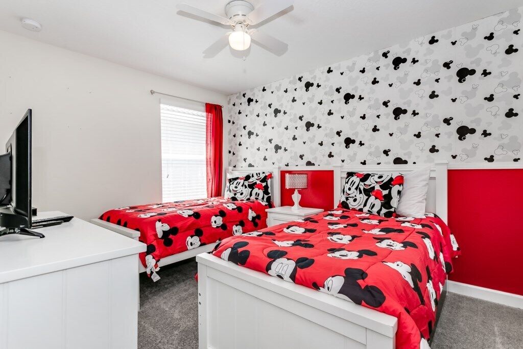 Ein Schlafzimmer mit zwei Betten, einem Schreibtisch, einem Deckenventilator und einer Wand mit Mickey Mouse Tapete.