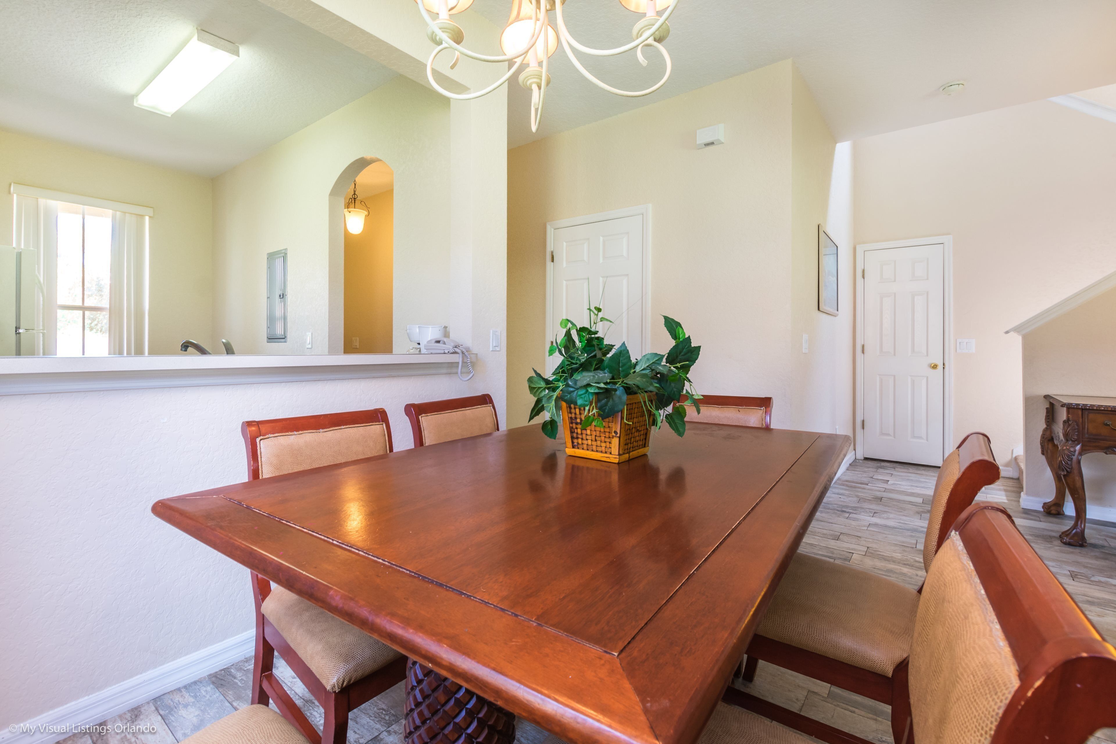 A dining room with a large wooden table, six chairs, a potted plant, and a chandelier.