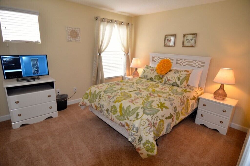 A bedroom with a bed, two bedside tables, a desk with a computer, a window with curtains, and wall art.