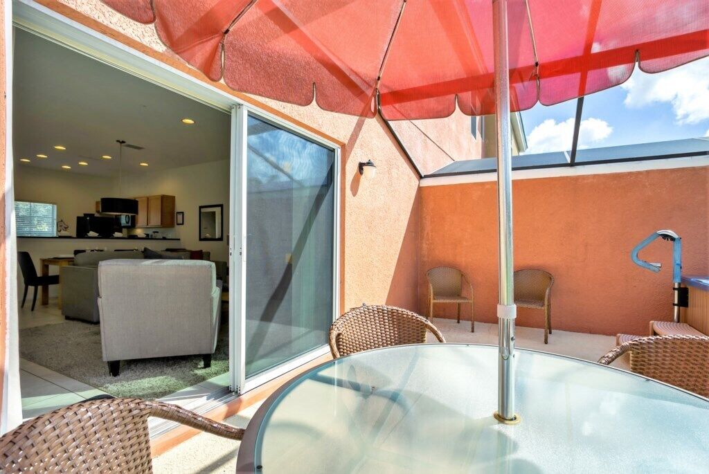 An outdoor patio with a glass table and wicker chairs, a red umbrella, and a sliding glass door leading to an interior living area with a sofa and kitchen appliances.