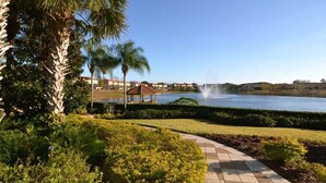 A landscaped area with a pathway, palm trees, and a body of water with a fountain.