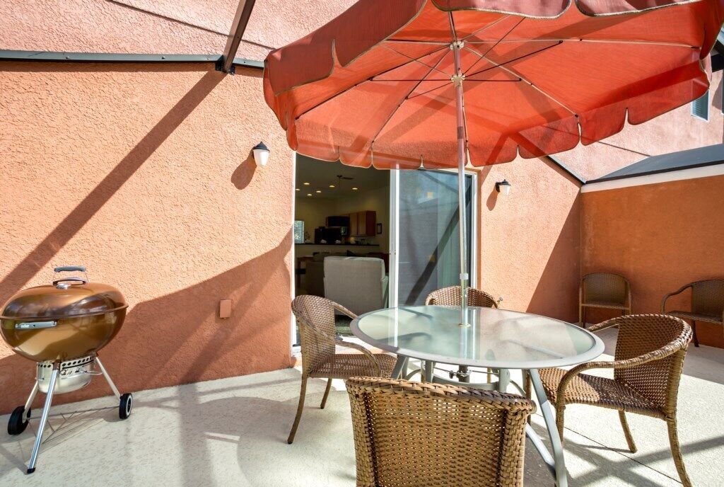 En udendørs terrasse med et rundt glasbord, fletstole, grill og en stor rød parasol.