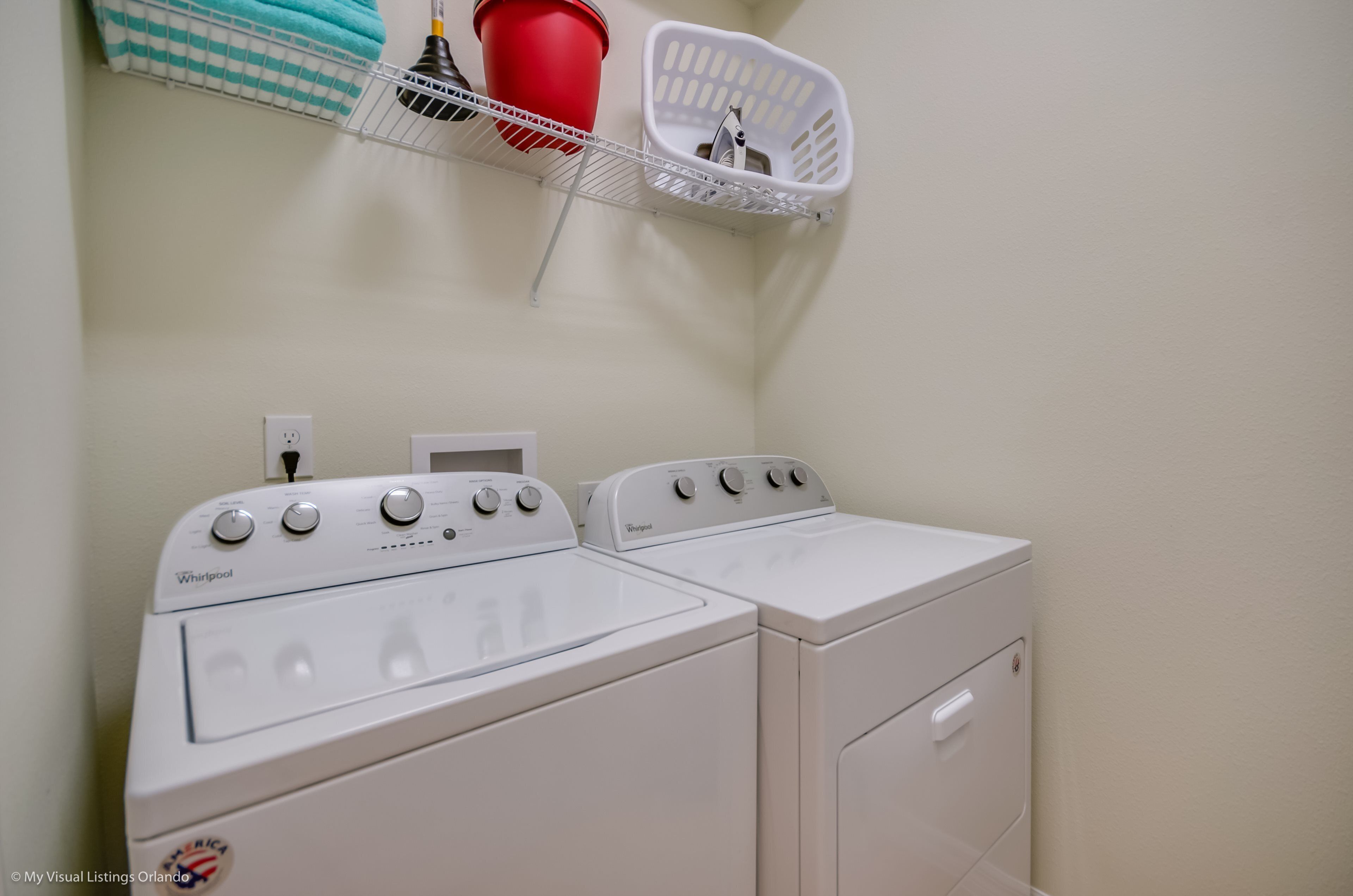 A laundry area with two white Whirlpool washers and dryers, a red bucket, and a white laundry basket on a wall-mounted shelf.