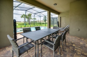 An outdoor patio with a table and chairs, a glass enclosure, and a view of a swimming pool and greenery.